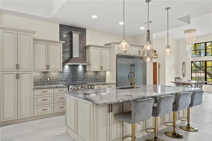 Kitchen with wall chimney exhaust hood, an island with sink, a kitchen breakfast bar, light stone countertops, and stainless steel built in refrigerator