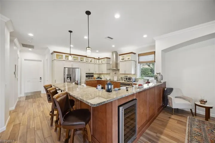 Kitchen with light stone counters, wall chimney range hood, stainless steel refrigerator with ice dispenser, beverage cooler, and white cabinetry