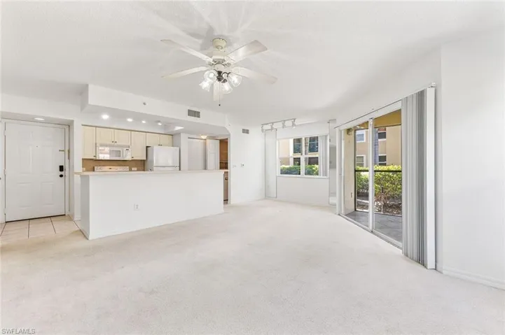 Spacious living area featuring light-toned carpeting, a ceiling fan with integrated lighting, and white baseboards, exterior slider to the lanai, and tile floors in the kitchen area.