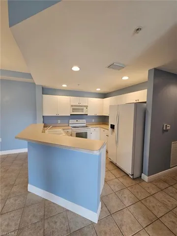 Kitchen with light tile flooring, kitchen peninsula, white cabinets, sink, and white appliances