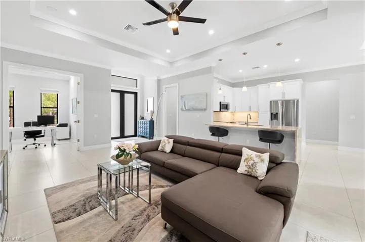 This expansive living area features light-colored tile flooring, a tray ceiling with recessed lighting and a ceiling fan, and an open layout that incorporates the kitchen