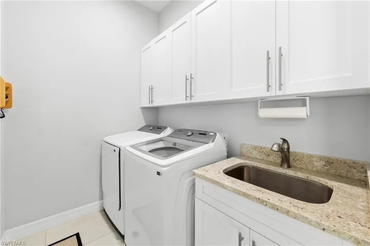 This utility space features a sink with a stone countertop, white cabinetry with silver hardware, and tile flooring