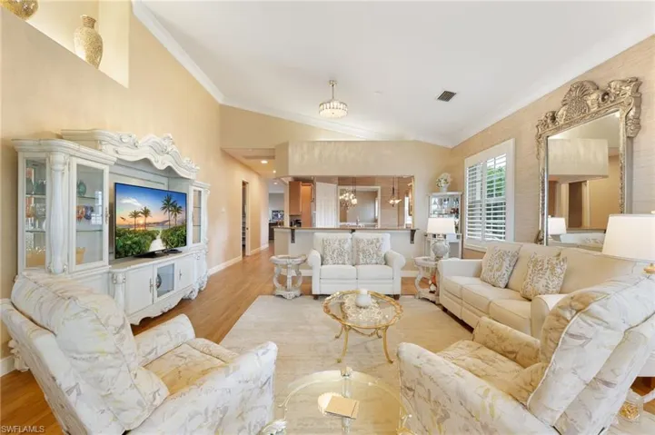 Spacious living area featuring vaulted ceilings, wood-finish flooring, plantation shutters, a decorative mirror, and a pass-through opening to an additional room