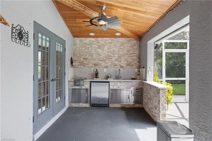 Outdoor summer kitchen featuring stacked stone accent walls, integrated stainless steel appliances, and a wood-finish vaulted ceiling with a ceiling fan