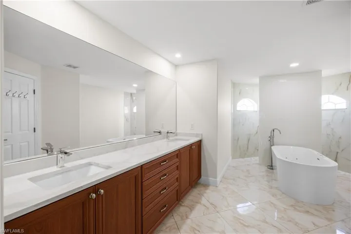 Spacious bathroom featuring a dual vanity with light wood cabinetry and white countertops, a large rectangular mirror, and a freestanding soaking tub with floor-mounted fixtures