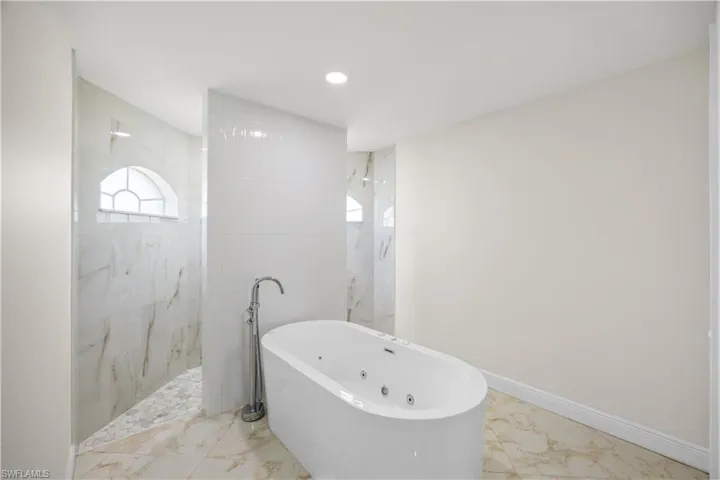 Freestanding soaking tub with chrome floor-mounted faucet, complemented by a walk-in shower featuring a half-moon window and marble-look tile walls