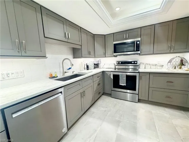 Kitchen featuring gray cabinetry, stainless steel appliances, light stone counters, crown molding, and a raised ceiling