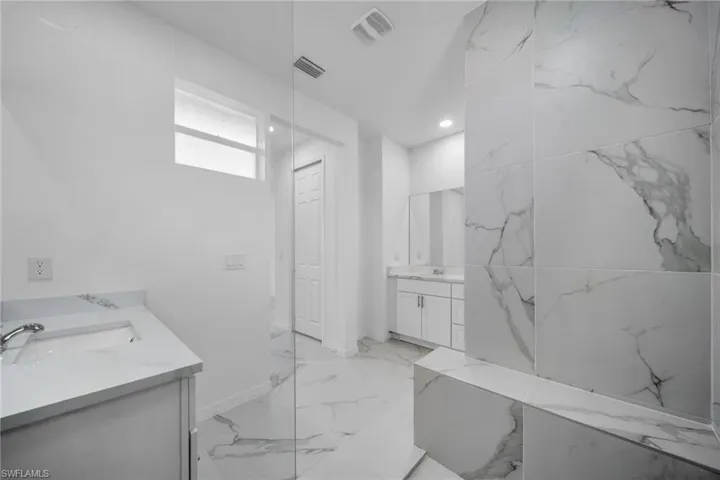 Spacious bathroom featuring large format marble-look tile flooring and shower surround, dual vanity areas with white cabinetry and integrated sinks, a clear glass shower partition, and a built-in shower bench