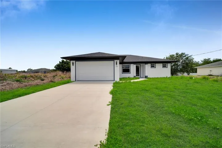 Contemporary single-story residence featuring a two-car garage, dark shingle roof, white exterior walls, a concrete driveway, and a vibrant green lawn