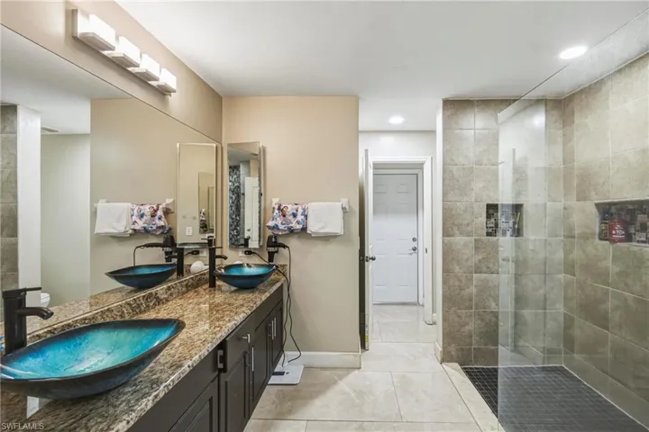 Spacious bathroom featuring a dual vanity with vessel sinks, a full-width mirror, a tiled walk-in shower with glass enclosure, recessed lighting, and tile flooring