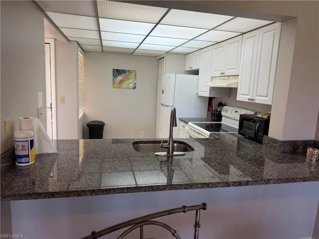 Kitchen featuring a peninsula, white cabinetry, white appliances, a paneled ceiling, and dark stone countertops