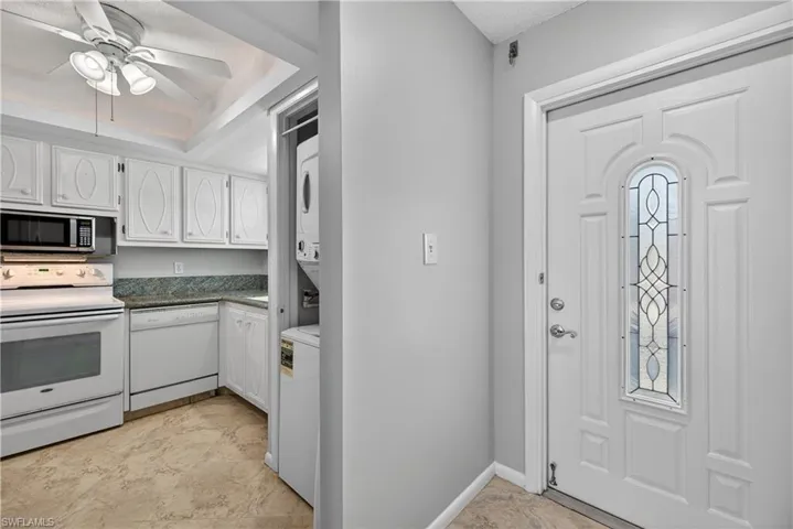 Entry door and kitchen featuring white cabinets, a ceiling fan/light, a tray ceiling with cove lighting, a stacked washer and dryer.