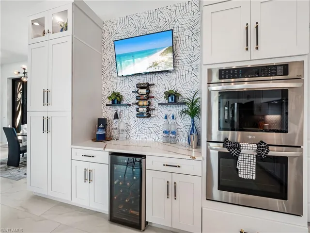 Bar with stainless steel double oven, wine cooler, white cabinetry, light stone counters, and glass fronted cabinets