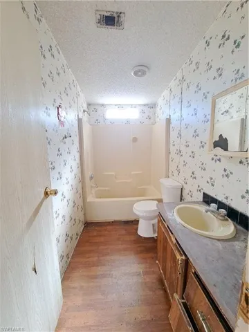 Bathroom with a textured ceiling, vanity, wallpapered walls, bathing tub / shower combination, and dark wood-type flooring