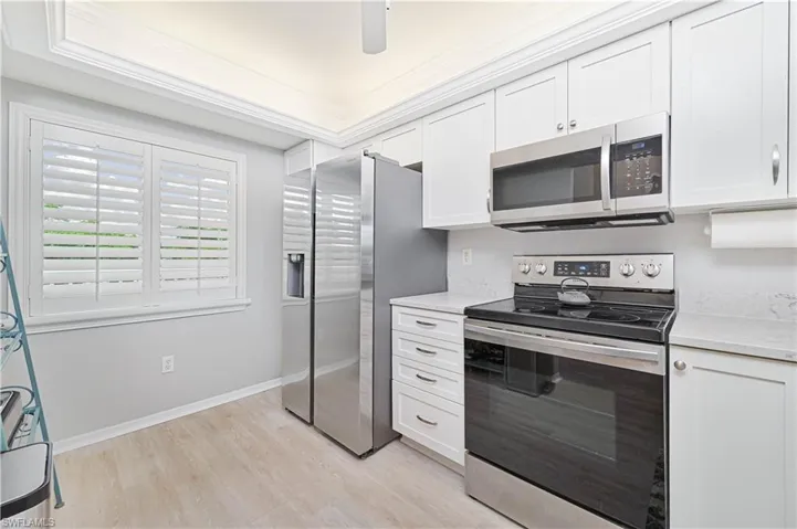 Kitchen with stainless steel appliances, plantation shutters, white cabinets, light stone counters, light wood-style flooring, and crown molding