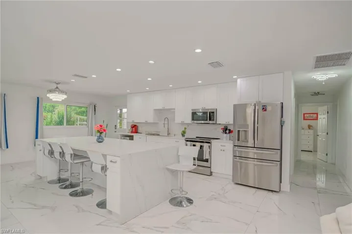 Spacious kitchen featuring an extended waterfall island, white cabinetry, stainless steel appliances, recessed lighting, and large format marble-look tile flooring