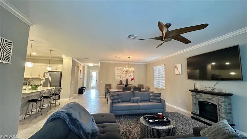 Living area with crown molding, light tile patterned floors, visible vents, a stone fireplace, and ceiling fan with notable chandelier