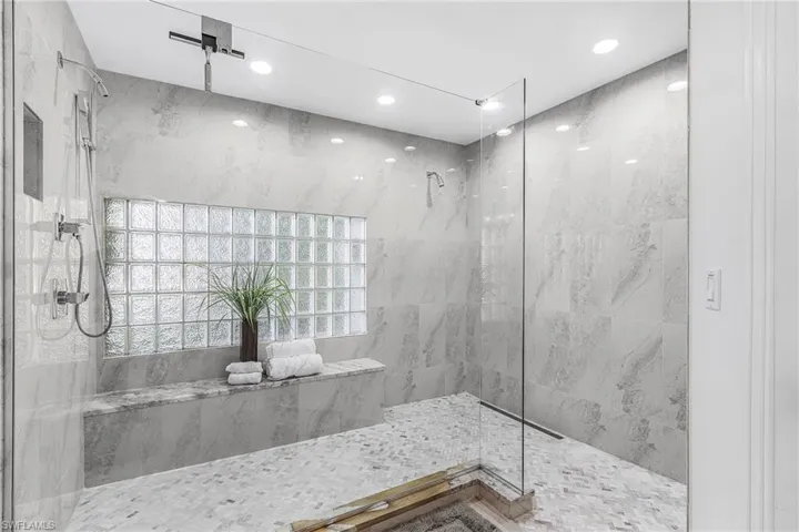 Spacious walk-in owners shower featuring floor-to-ceiling marbled tile, a built-in bench, a glass block window, dual shower heads, and recessed lighting