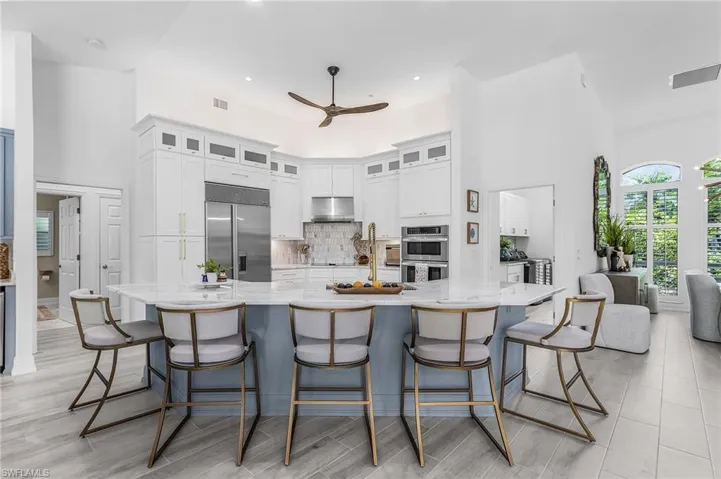 Expansive kitchen featuring a large central island with seating for five, white cabinetry with upper glass-front accents, stainless steel appliances, and a tiled backsplash