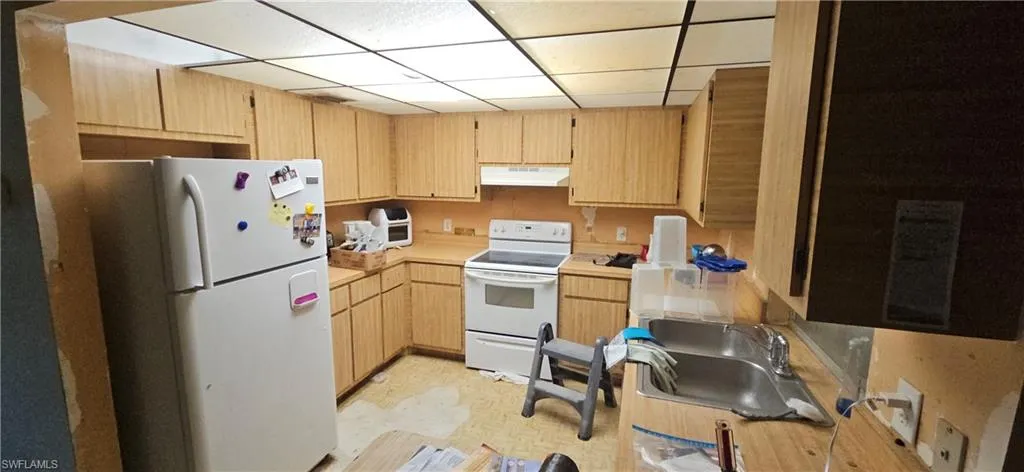 Kitchen with white appliances, light brown cabinetry, sink, and a drop ceiling