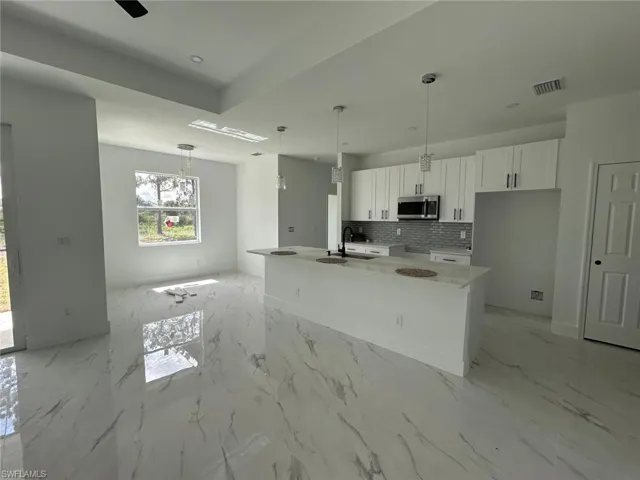Kitchen with stainless steel microwave, white cabinets, backsplash, a center island with sink, and light marble finish flooring