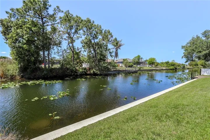 Waterfront property featuring a concrete seawall, lush green lawn, mature trees, and a spacious canal with floating lily pads