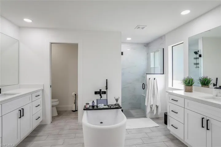 Expansive bathroom featuring a freestanding soaking tub, dual vanity with white cabinetry and granite countertops, and a glass-enclosed shower with tiled walls and matte black fixtures