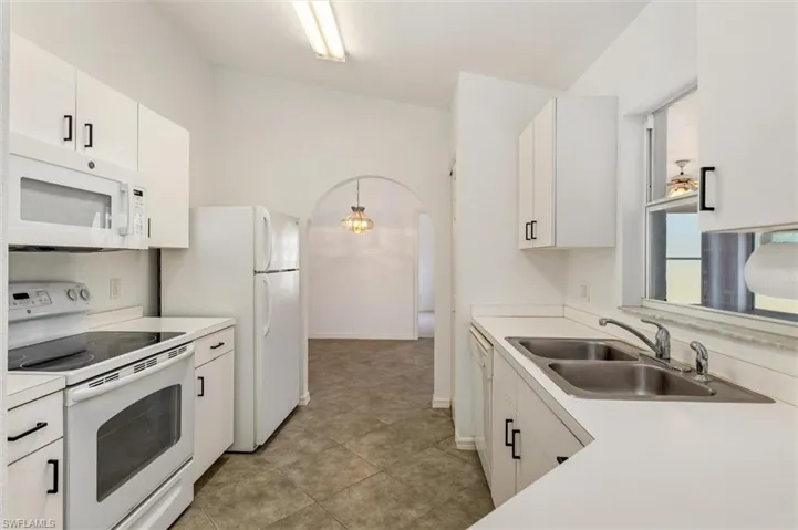 Galley kitchen featuring white cabinetry with black hardware, white appliances, a double basin stainless steel sink, and tile flooring
