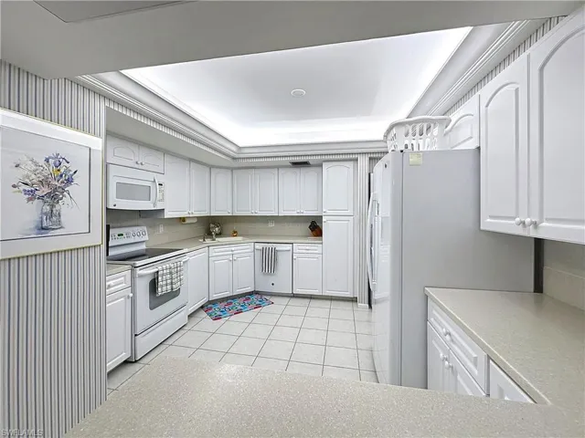 Kitchen with a sink, light tile patterned flooring, white appliances, a raised ceiling, and white cabinetry