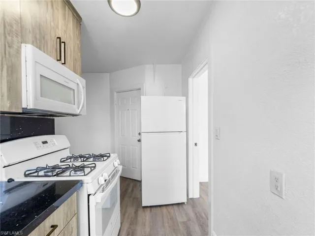 Kitchen with white appliances, light wood-style flooring, a textured wall, and dark stone counters