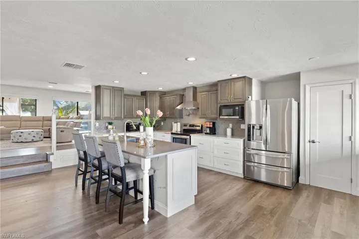 Kitchen with a breakfast bar, wood-type flooring, wall chimney range hood, stainless steel appliances, and a center island with sink