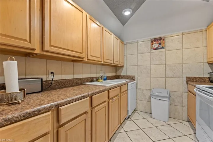 Kitchen featuring white appliances, a sink, light brown cabinetry, dark countertops, and vaulted ceiling