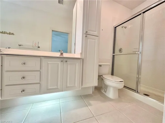 Bathroom featuring a white vanity with under-cabinet lighting, a frameless mirror, a toilet, a glass-enclosed shower, and light-toned floor tiling