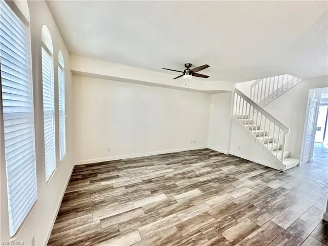 Spacious living area featuring wood-look tile flooring, three arched windows with horizontal blinds, a ceiling fan, and a carpeted staircase with white railings