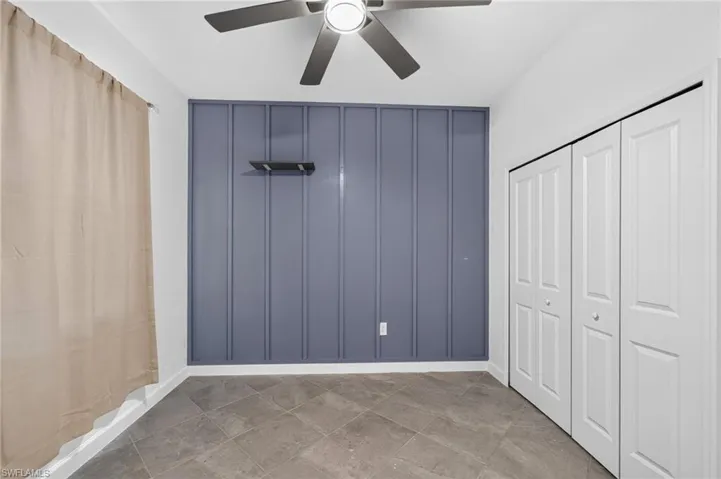 Room featuring a vertical-panel accent wall, dark-toned tile flooring, dual bi-fold closet doors, and a ceiling fan with integrated lighting