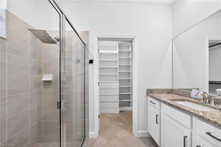 Bathroom featuring a walk-in shower with glass enclosure, wall-mounted rainfall showerhead, stone-finish countertop with undermount sink, white cabinetry with dark hardware, and built-in closet shelving