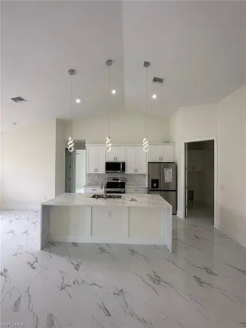 Kitchen featuring light stone counters, hanging light fixtures, stainless steel appliances, white cabinetry, and a kitchen island with sink