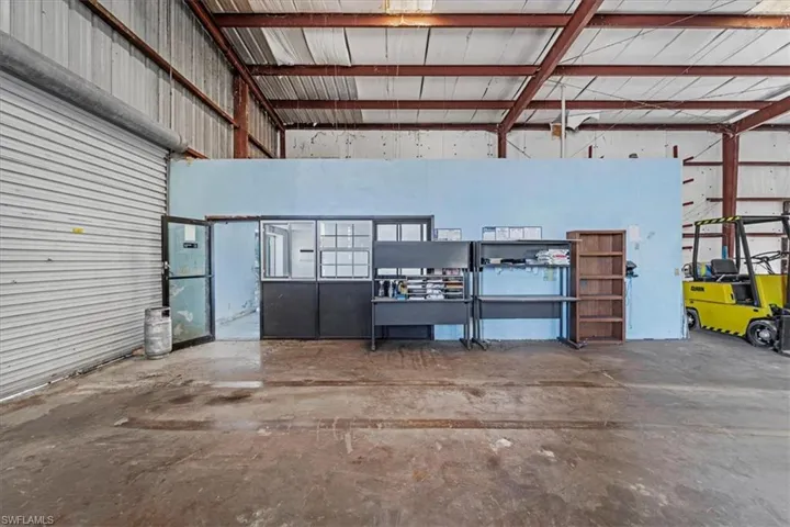 Industrial warehouse space featuring a large roll-up door, exposed structural beams, a concrete floor, and integrated office partitioning with a glass-paneled entry door