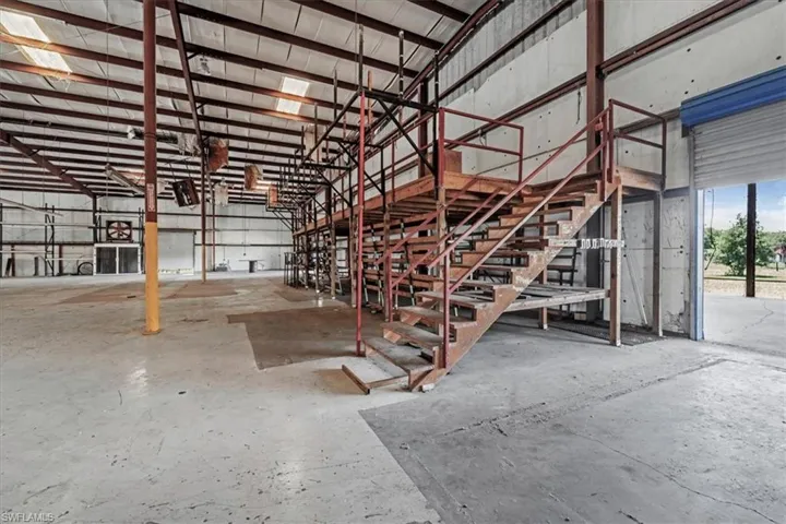 Spacious warehouse interior featuring a metal mezzanine with an industrial staircase, exposed steel beams, concrete flooring, and a large roll-up door