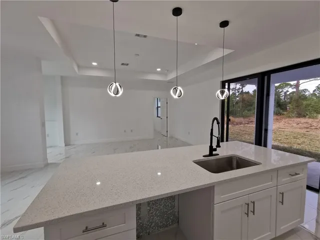 Kitchen featuring light marble finish floors, pendant lighting, light stone counters, a kitchen island with sink, and a raised ceiling