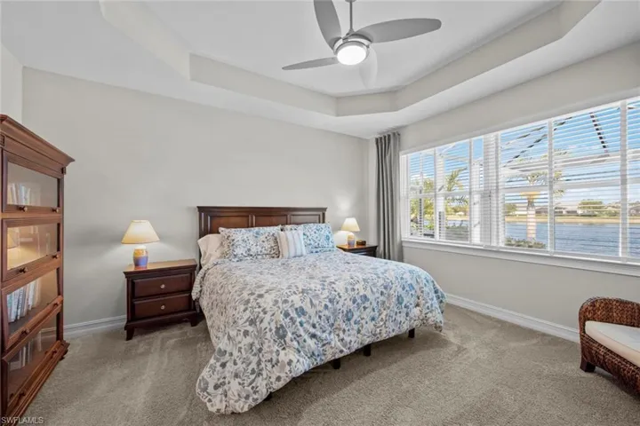 The large primary bedroom features views of the lake, TRAY CEILING, floor to ceiling linen curtains (September 2021), window blinds (April 2017), and ceiling fan/light (November 2020).