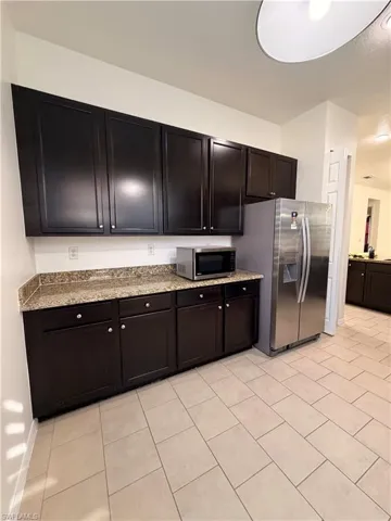 Dark wood-finish cabinetry, light-toned tiled flooring, stone-finish countertops, and stainless steel refrigerator