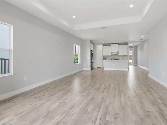 Spacious open-concept living area featuring light wood-finish flooring, a contemporary kitchen with white cabinetry, stainless steel appliances, and a tiled backsplash