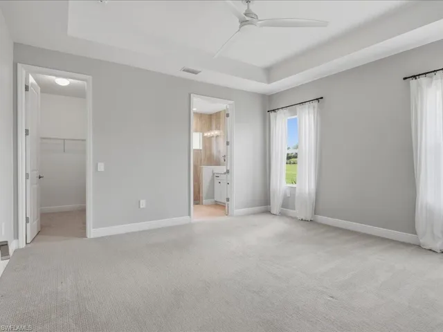 Spacious room featuring light gray carpeting, a tray ceiling with a modern ceiling fan, and multiple windows with white sheers