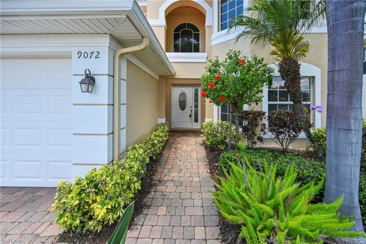 Stucco exterior with arched entry, paver walkway, white paneled garage door, and lush landscaping