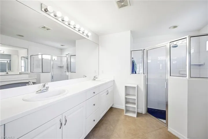 Dual vanity with a white countertop and integrated sinks, large wall-mounted mirror, brushed nickel fixtures, glass-enclosed shower, and neutral tile flooring