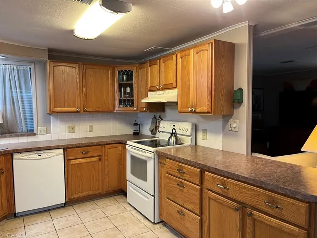 Kitchen with brown cabinets, dark countertops, glass insert cabinets, white appliances, and under cabinet range hood