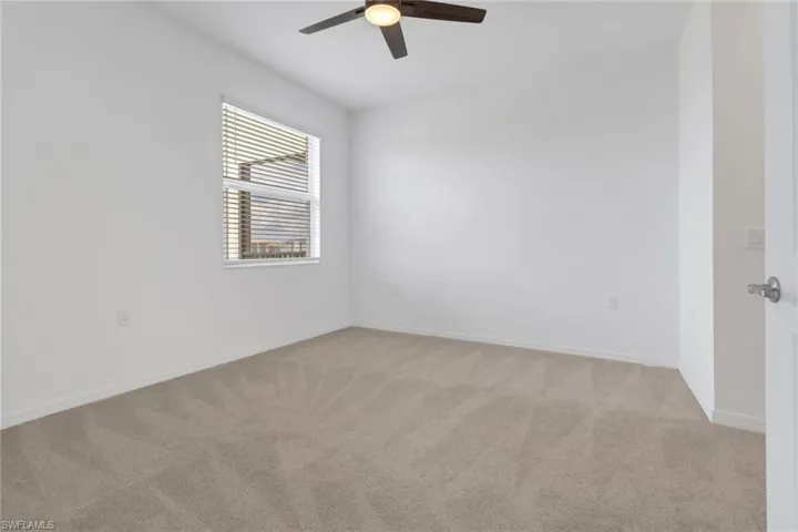 Spacious room featuring neutral carpeting, white walls, a single window with blinds, a ceiling fan with light fixture, and white trim