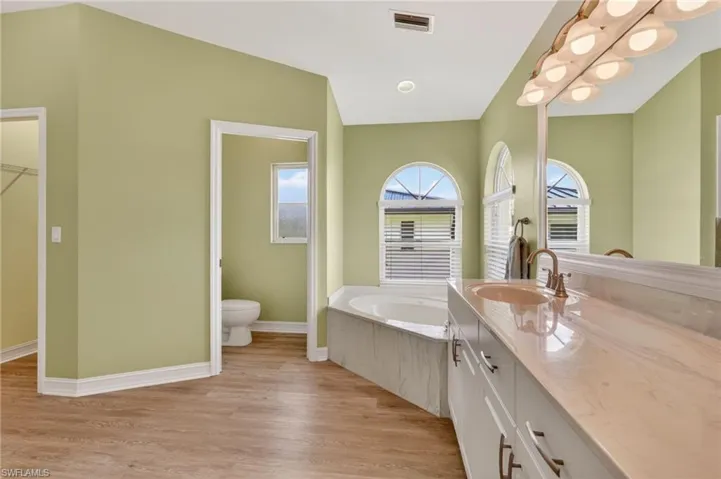 Bathroom featuring a double vanity with an integrated sink and large mirror, a corner soaking tub, two arched windows with blinds, a separate water closet, and wood-finish flooring