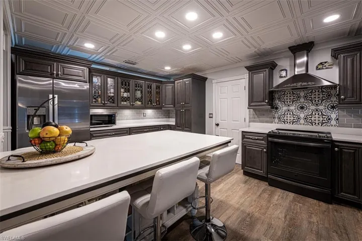 Kitchen with backsplash, dark wood-type flooring, wall chimney exhaust hood, appliances with stainless steel finishes, and dark brown cabinets